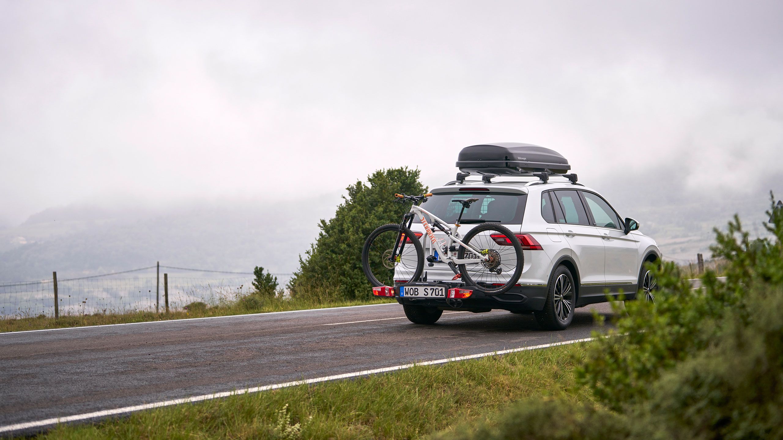 VW Tiguan so strešným boxom a nosičom bicyklov z originálneho príslušenstva Volkswagen vrátane bicykla na poľnej ceste