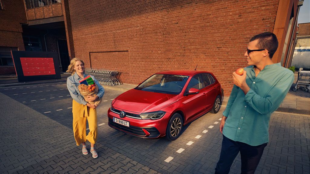 Žena s taškou plnou potravín stojí pri zaparkovanom červenom aute. Muž vedľa nej, ktorý drží jablko, sa pozerá jej smerom. Obaja sú na parkovisku vedľa tehlovej budovy.