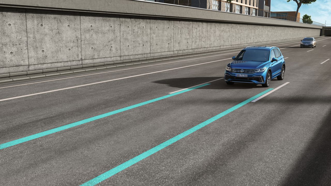 Na obrázku sú dve osobné vozidlá jazdiace po viacprúdovej ceste pri betónovej stene. Na vozovke sú vyznačené dva modré pruhy systému zmeny jazdného pruhu. Prvé auto je modré a druhé strieborné.