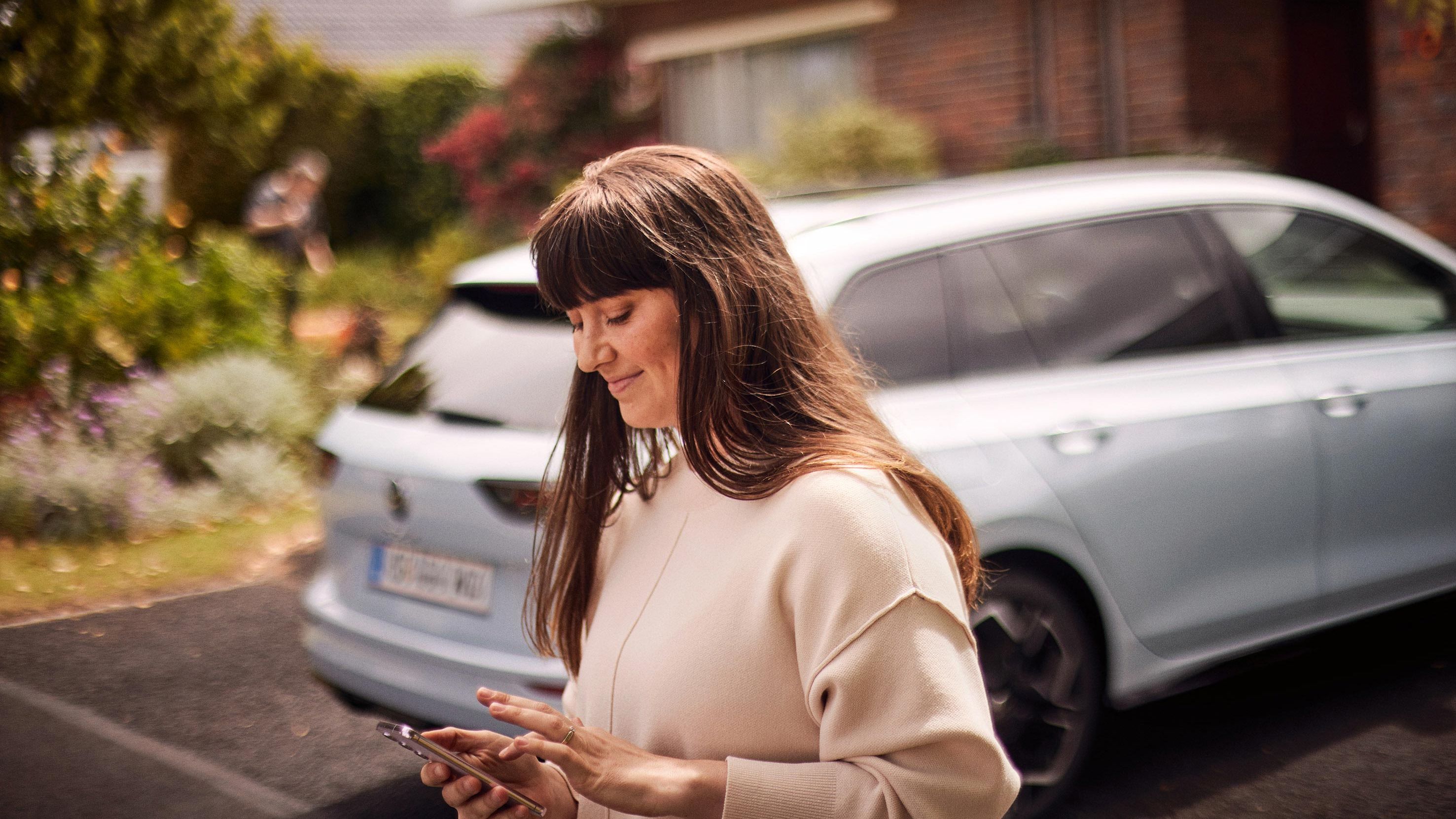 Žena stojí za vozidlom Volkswagen a pozerá na svoj smartfón, ktorým niečo ovláda. Vozidlo je rozostrené a stojí pred domom. Za vozidlom je krásna záhrada.