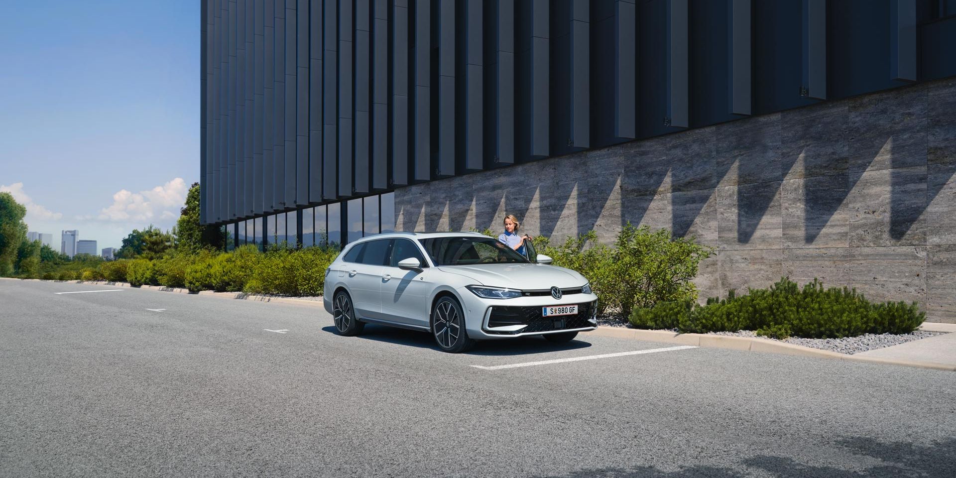 Biele auto zaparkované pred modernou budovou s tmavou fasádou. Žena nastupuje do vozidla na mieste vodiča. Vedľa budovy je zeleň.