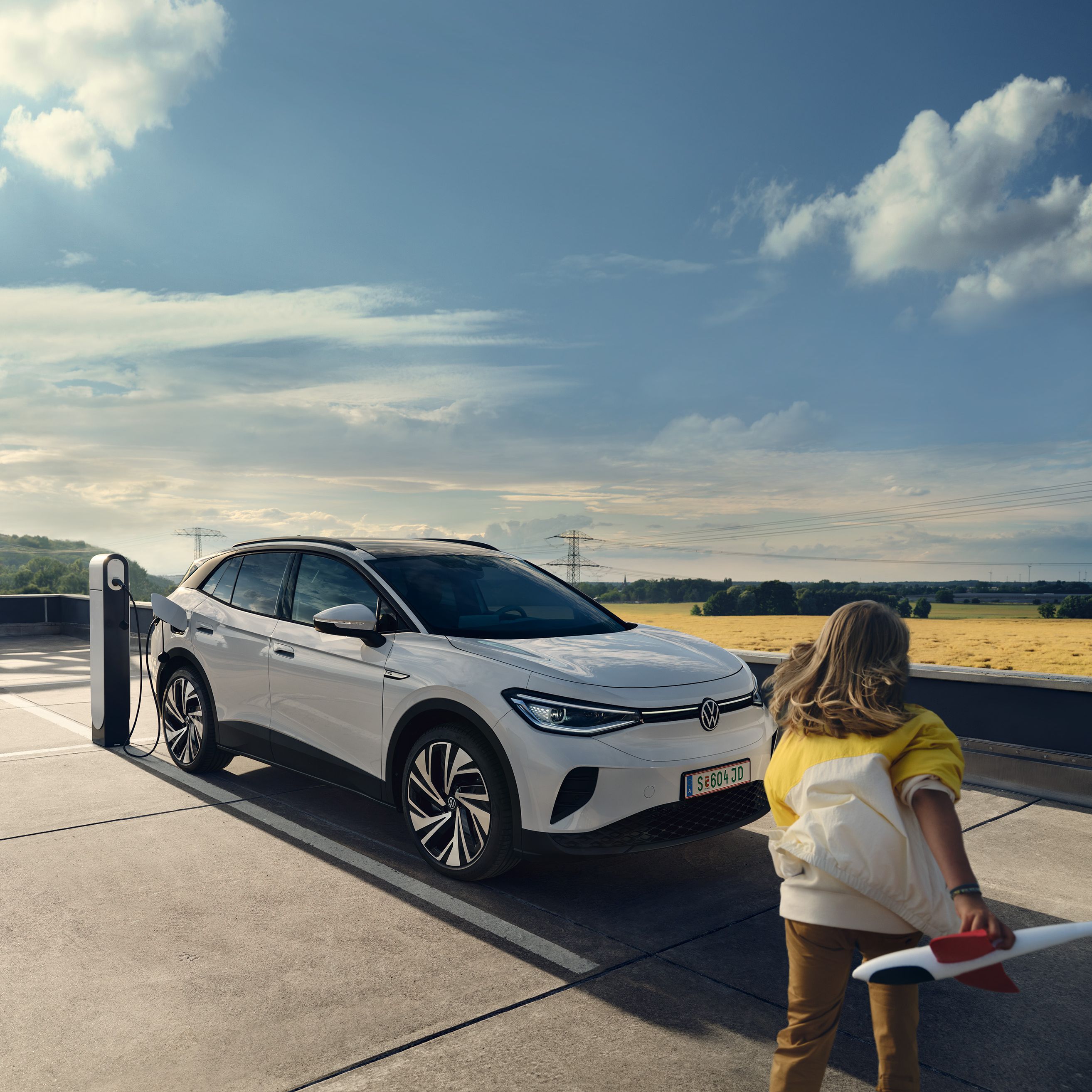 Žena drží model lietadla a prichádza k bielemu elektrickému SUV, ktoré sa nabíja na nabíjacej stanici pod modrou oblohou. Auto je zaparkované na vonkajšom parkovisku. V pozadí sa rozprestiera lúka a niekoľko stromov.