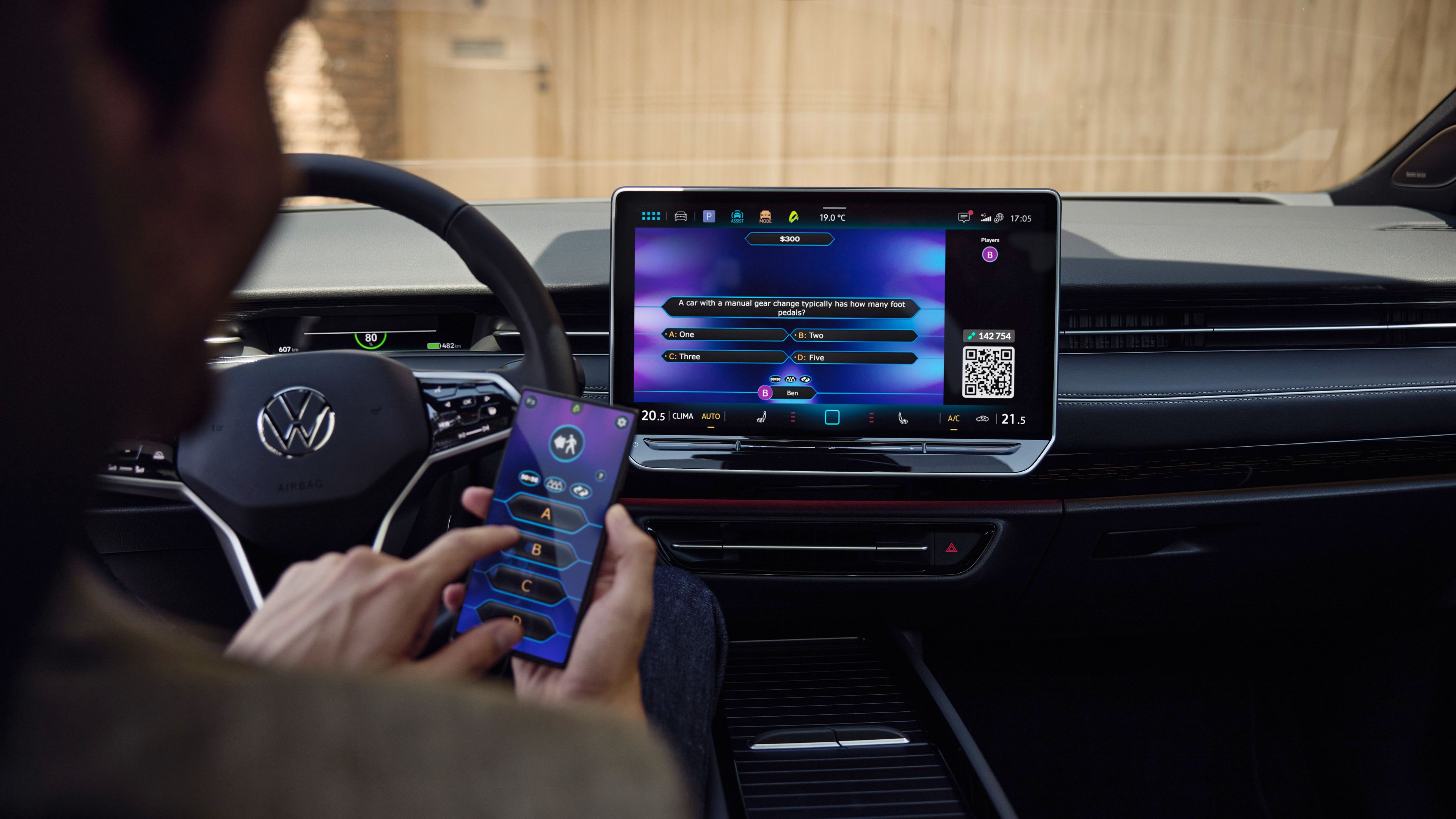 Muž sedí v aute a drží mobilný telefón, na ktorom je zobrazená aplikácia. Na palubnej doske pred ním je veľká obrazovka s podobnou aplikáciou. Ide o moderný interiér vozidla Volkswagen.
