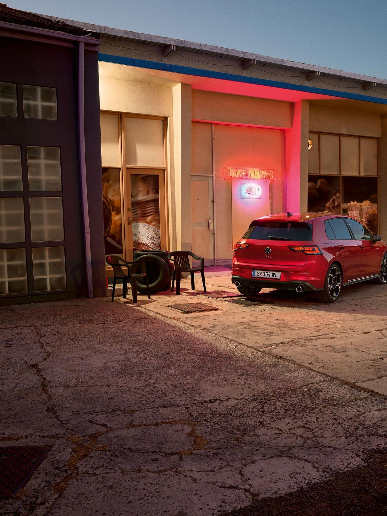 Červený Golf GTI je zaparkovaný pred svetlou budovou s nápisom "SHOP" na fasáde. Dve osoby kráčajú vedľa auta po betónovej ceste. Budova má osvetlený výklad a vedľa vchodu sú umiestnené stoličky a pneumatika. Za súmraku.
