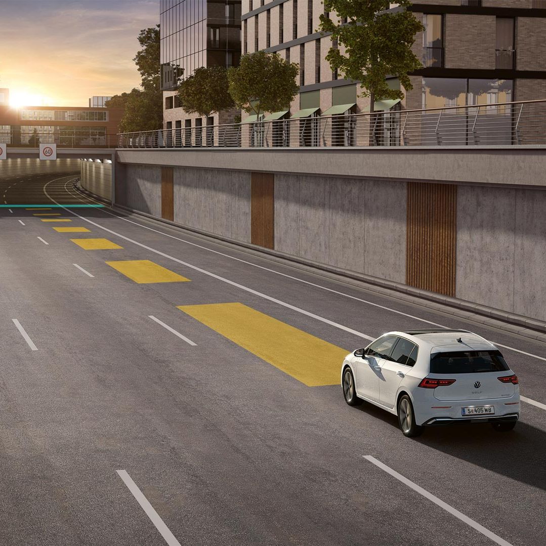 Biely VW Golf jazdí po ceste s aktívnym adaptívnym tempomatom ACC.  Vozidlo smeruje do tunela. V pozadí vidieť mesto pri zapadajúcom slnku.