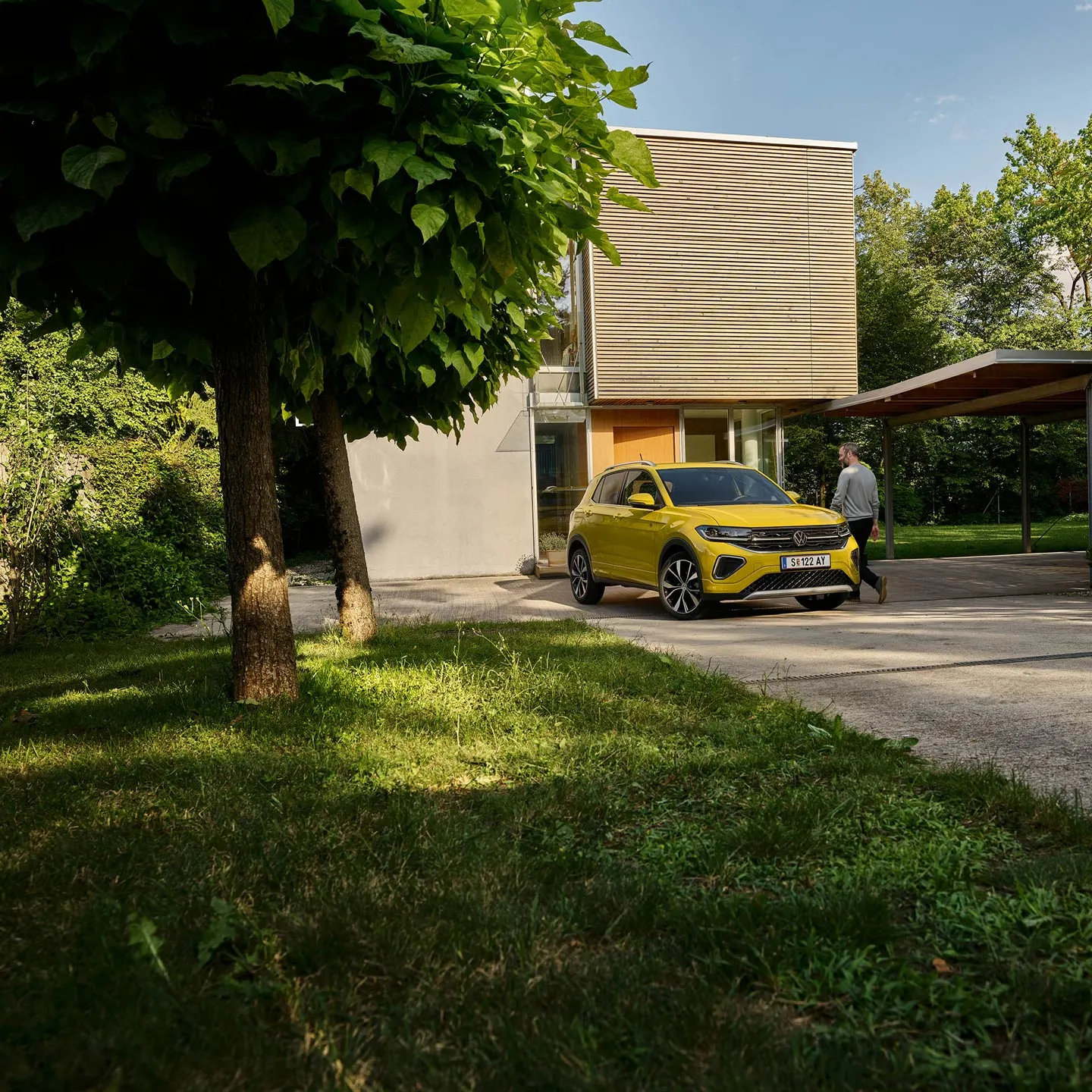 Muž sa chystá nastúpiť do žltého VW T-Cross, ktoré je zaparkované na príjazdovej ceste k domu.