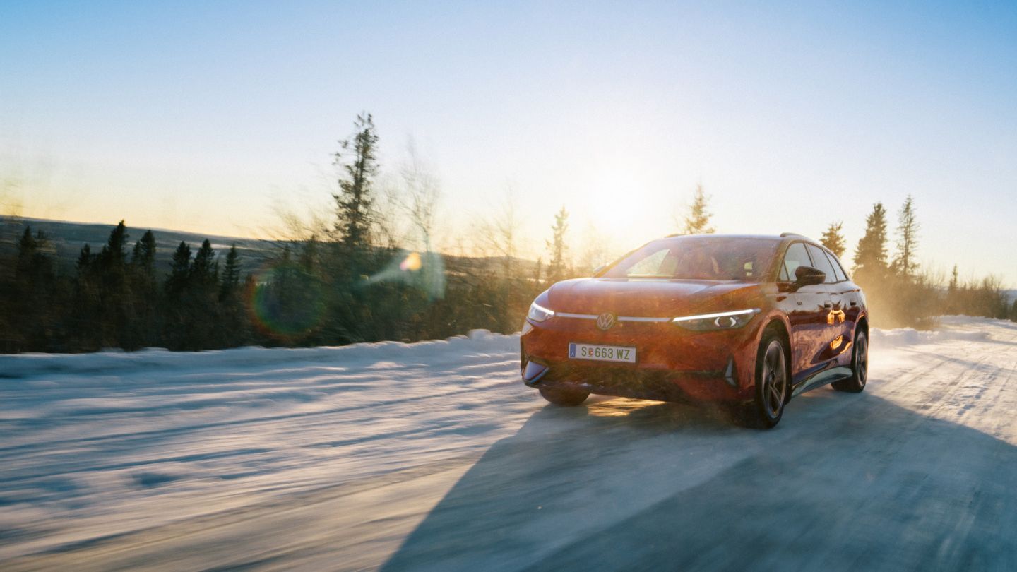 Červený VW ID.7 Tourer GTX jazdí po vidieckej ceste pri západe slnka. Zapadajúce slnko vrhá teplé, zlatisté svetlo na okolie aj na vozidlo.