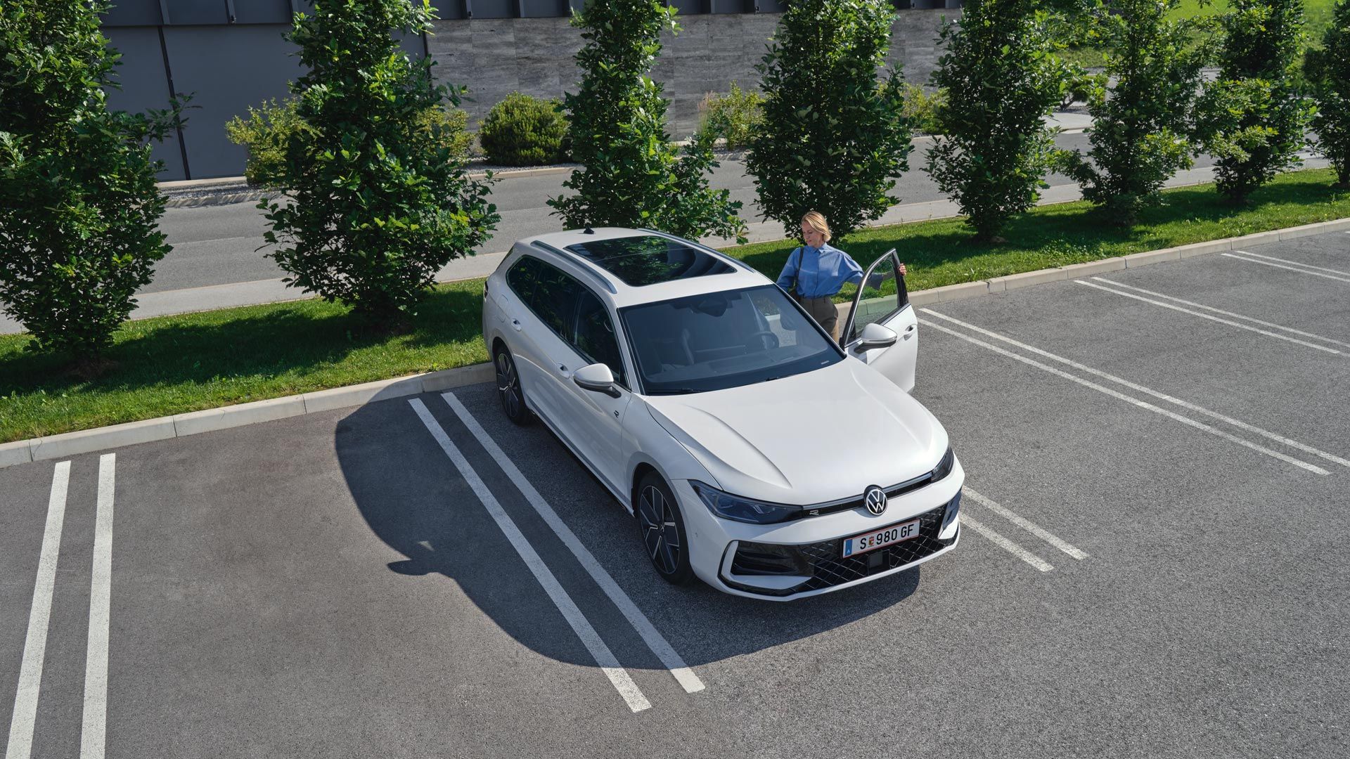 Pohľad zhora na biely VW Passat na parkovisku so ženou, ktorá nastupuje na strane vodiča.