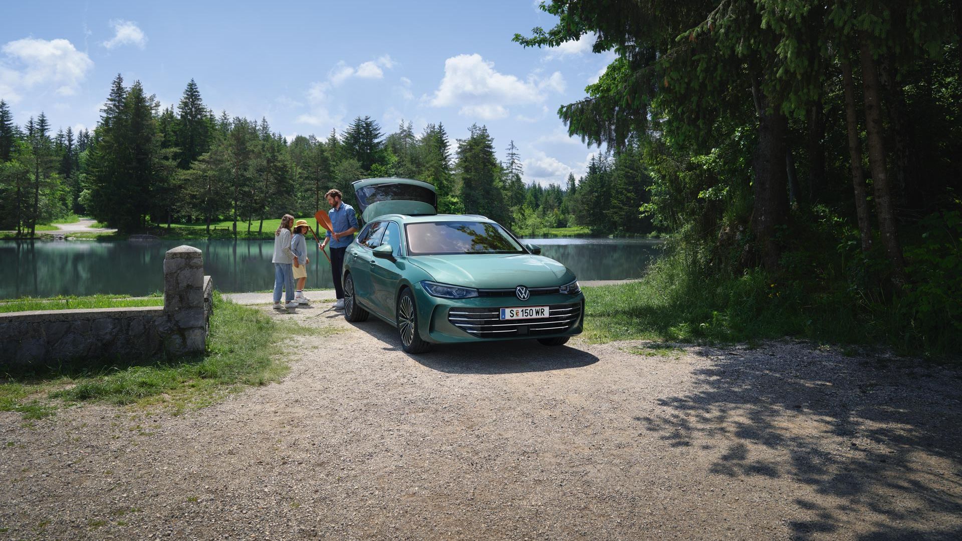 Modrozelený VW Passat s otvoreným batožinovým priestorom parkuje pri jazere. Muž rozdáva hračky dvom deťom. Okolo jazera je veľa zelene a stromov.