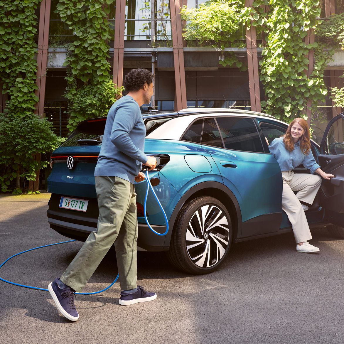 Muž s nabíjacím káblom v ruke kráča k modrému VW ID.4 zaparkovanému na dvore.
