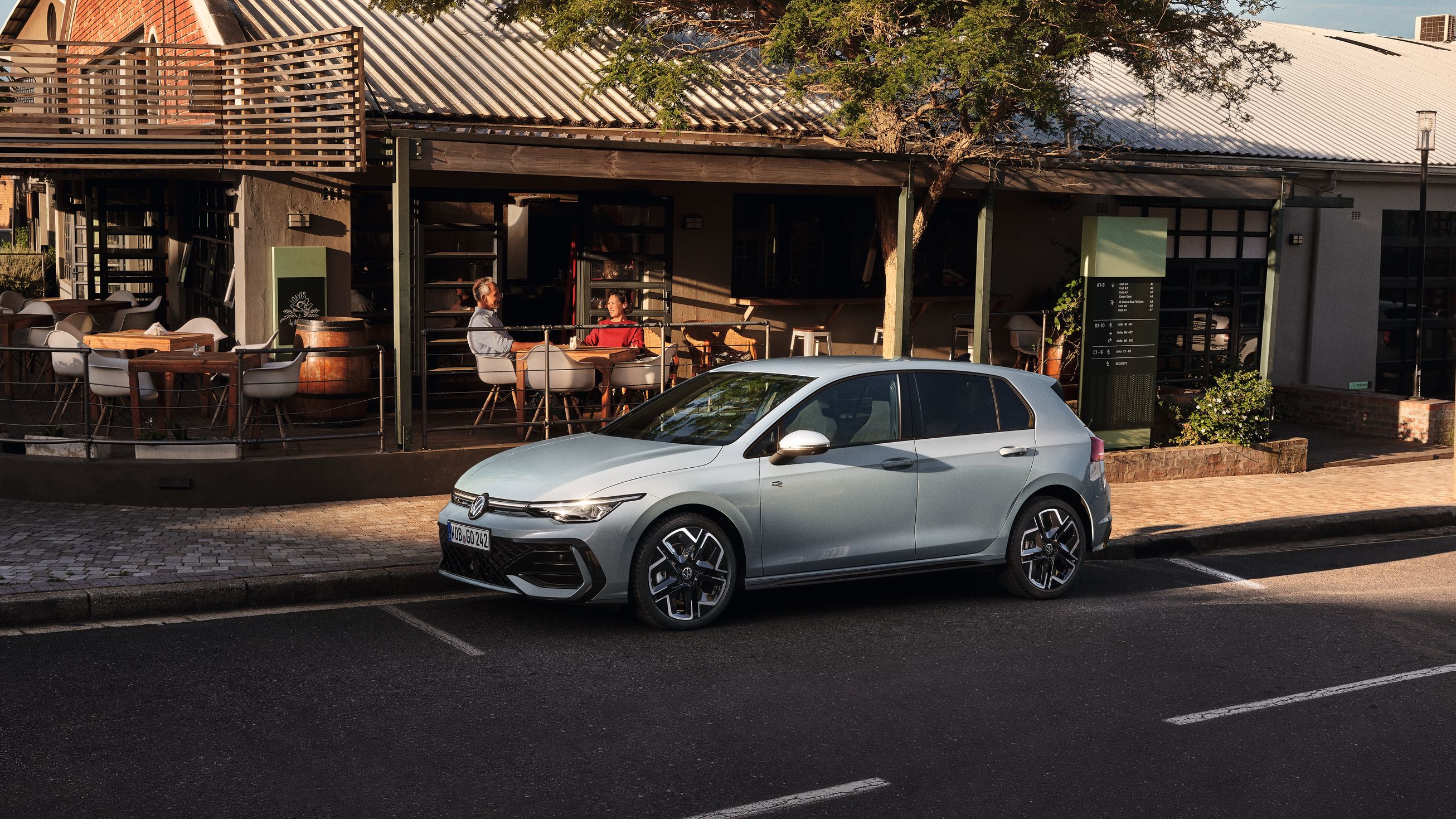 Biely Golf R-Line zaparkovaný na parkovacom mieste na ceste pred reštauráciou. Za vozidlom je reštaurácia a na terase sedia zákazníci.