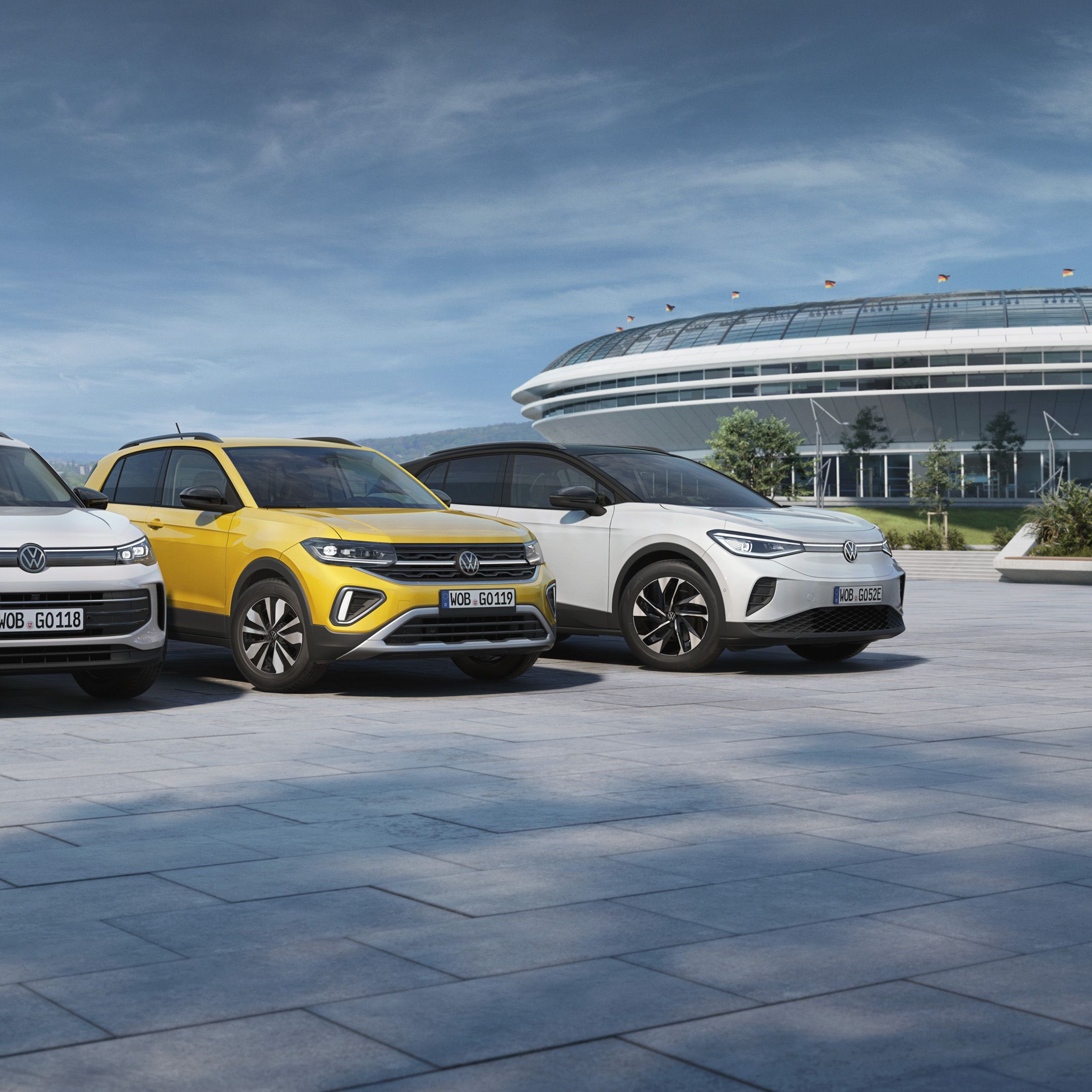 Tri vozidlá Volkswagen stoja na ploche pred modernou multifunkčnou budovou, v pozadí vidieť mesto a hory.
