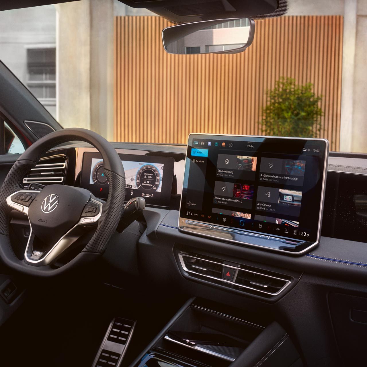 Pohľad do interiéru VW Tiguan zameraný na volant, digitálny prístrojový panel, displej systému infotainment, palubnú dosku, časť stredovej konzoly a výhľad z čelného skla na modernú stavbu v meste.