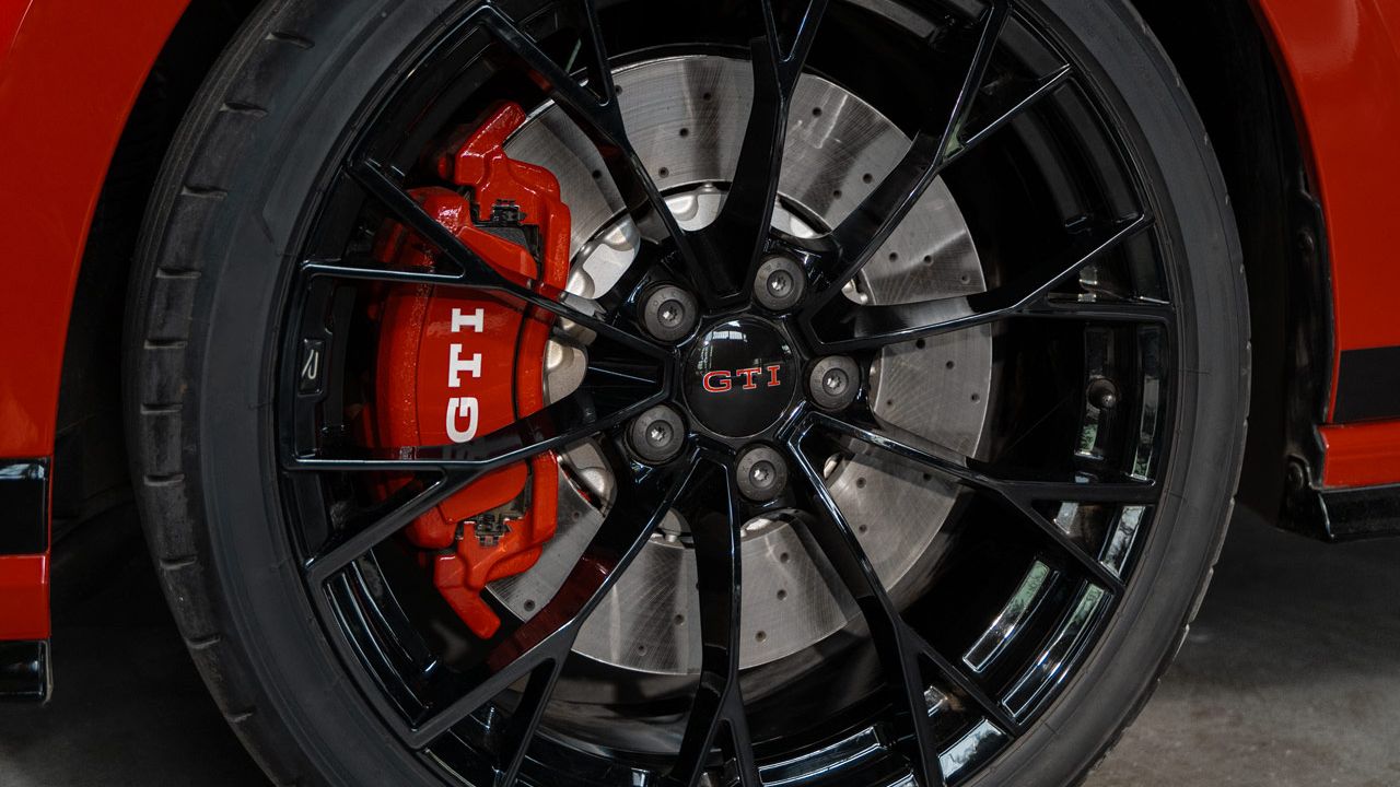 Detailný záber červeného Golfu GTI s čiernymi kolesami. Na brzdovom strmeni je viditeľné logo GTI. Vozidlo je zaparkované v priemyselnom prostredí.
