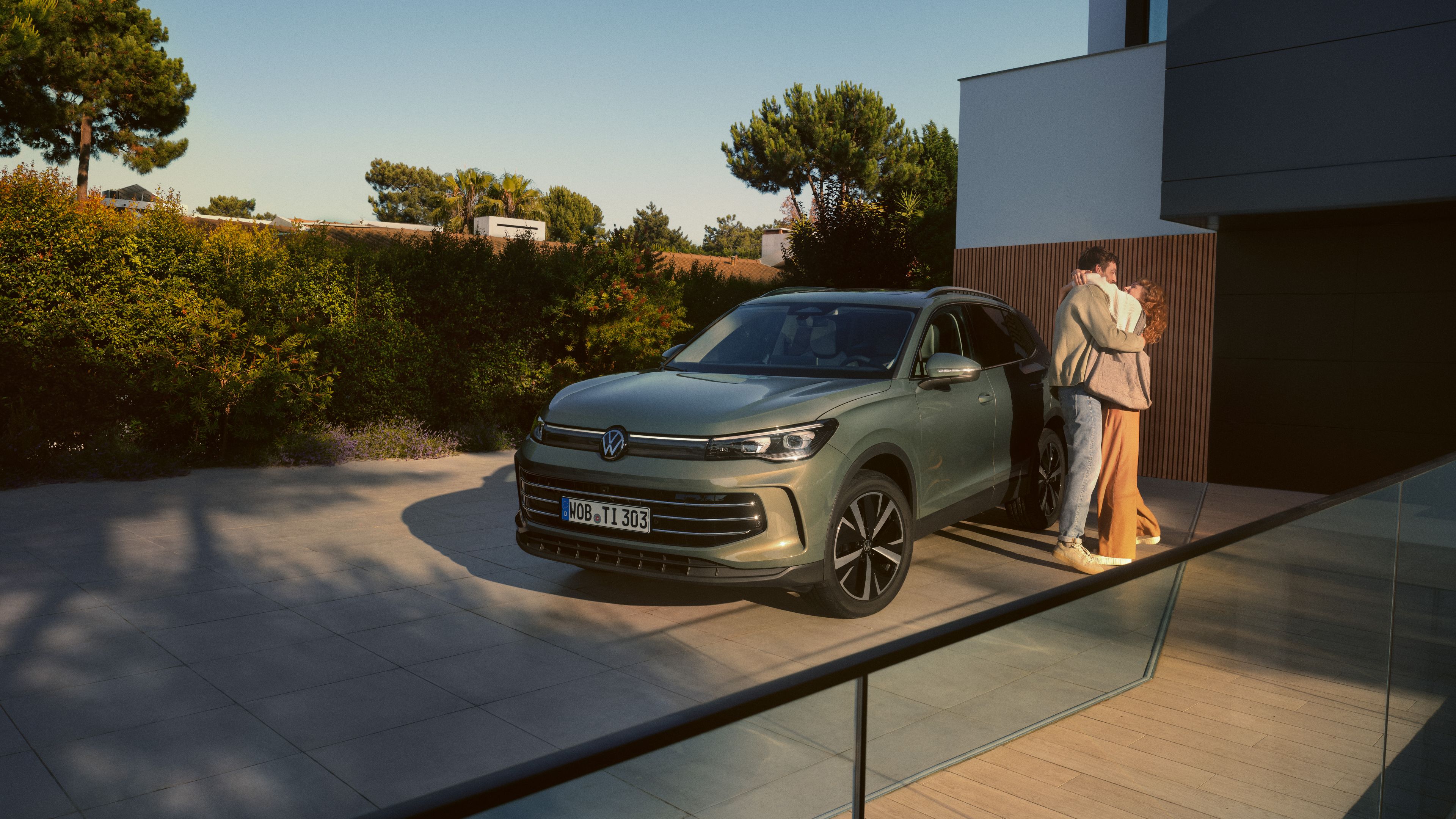Zelený VW Tiguan zaparkovaný pre moderným domom.dernen Wohnhauses. Ein junges Paar steht daneben und umarmt sich.
