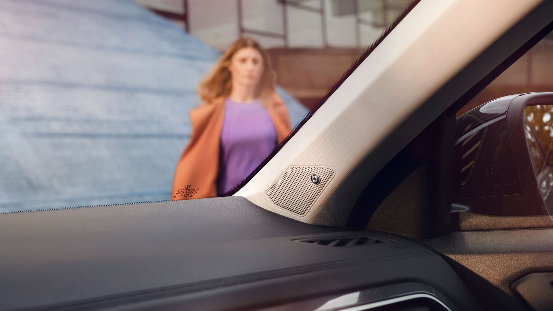 Interiér VW T-Roc, detail na reproduktor zvukového systému BeatsAudio™ integrovaného do A-stĺpiku. Cez čelné sklo vidieť ženu, ktorá kráča k vozidlu a za ňou budovu.