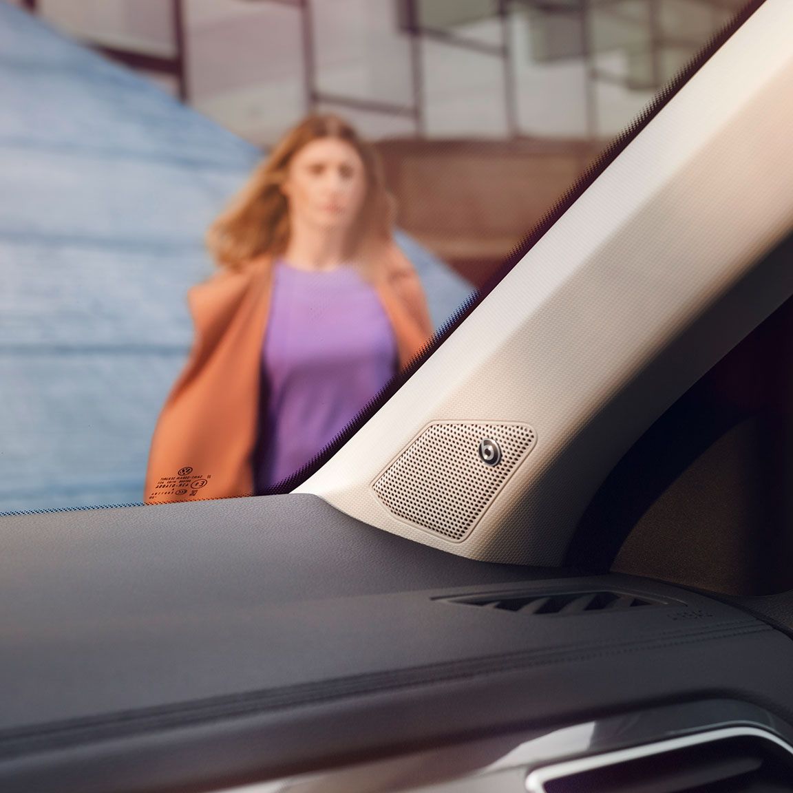 Interiér VW T-Roc, detail na reproduktor zvukového systému BeatsAudio™ integrovaného do A-stĺpiku. Cez čelné sklo vidieť ženu, ktorá kráča k vozidlu a za ňou budovu.