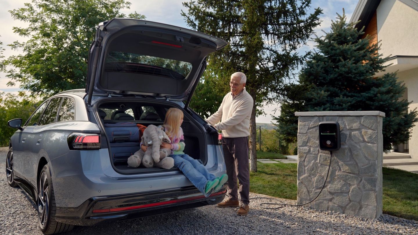 Ein älterer Mann lädt den VW ID.7 Tourer an der Wallbox, im Kofferraum sitzt ein Kind und wartet. 