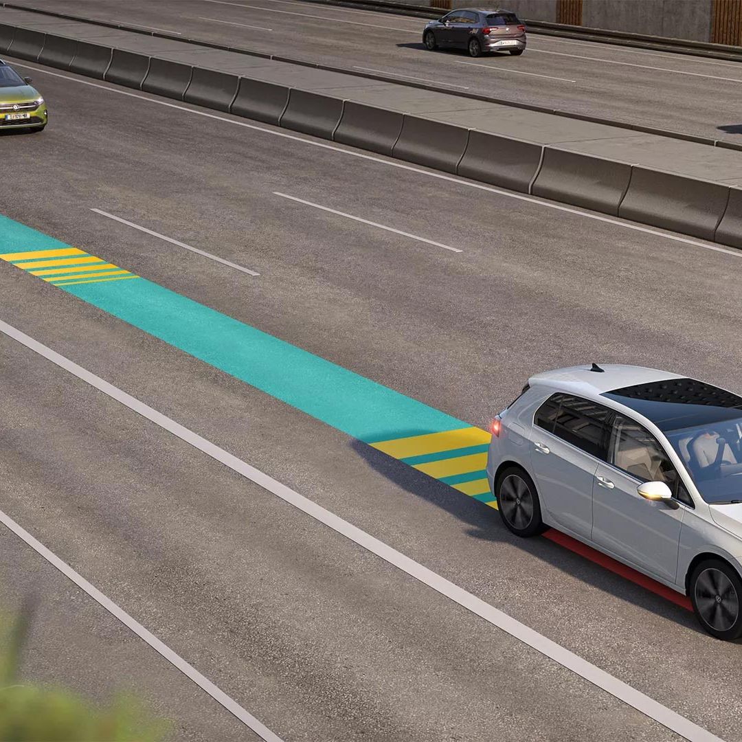 Biely VW Golf jazdí v premávke s aktívnym systémom Emergency Assist, schematicky znázornený na obrázku modro-žltým pásom.