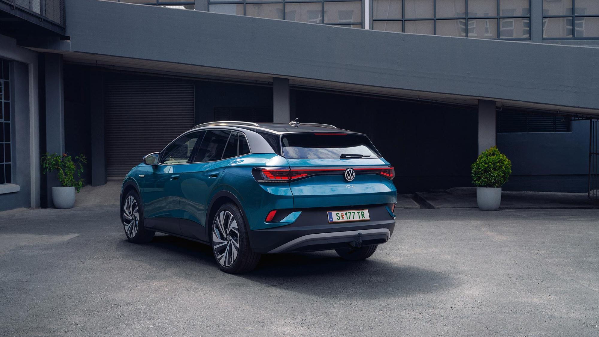 Modrý elektrický automobil Volkswagen ID.4 stojí zaparkovaný pred modernou industriálnou budovou s sklenenou fasádou. Má elegantné línie a športový vzhľad. Na okraji parkoviska sú dve zelené rastliny v kvetináčoch.
