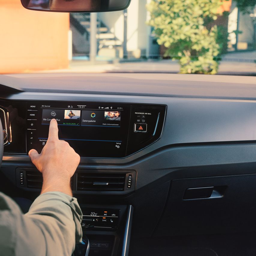 Muž ovláda internetový obchod VW upgrads vo VW Polo prostredníctvom displeja systému infotainment. Čiastočne vidieť volant a digitálny prístrojový panel. Cez čelné sklo vidieť strom a budovu.