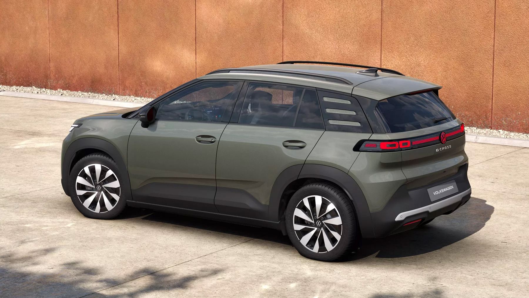 Olivovo zelený VW ID. Cross Style stojí na betónovej ploche pred hrdzavou stenou. Vozidlo má čierne strešné nosiče, tónované okná a charakteristické LED zadné svetlá s niekoľkými horizontálnymi pruhmi. Disky sú dvojfarebné v čiernej a striebornej farbe.