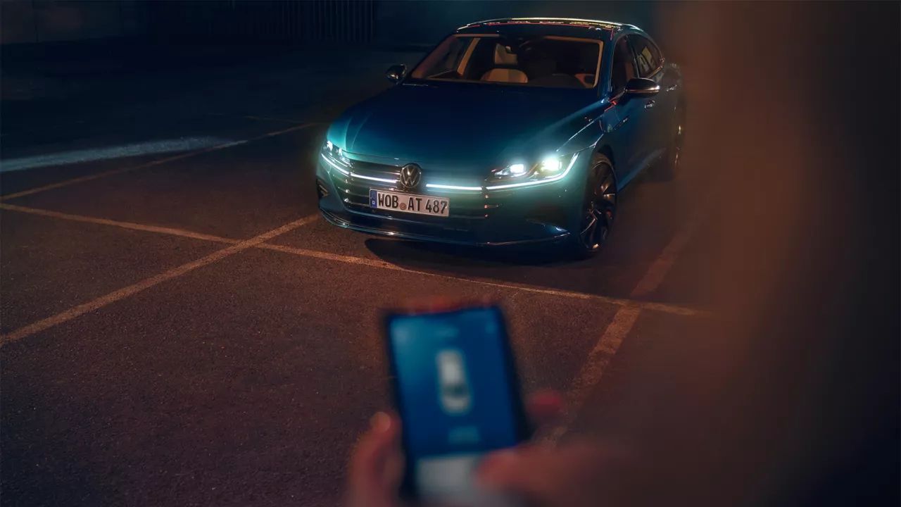 Osoba stojí pred vozidlom Volkswagen a drží smartfón v ruke. Na displeji je zobrazená aplikácia Volkswagen.