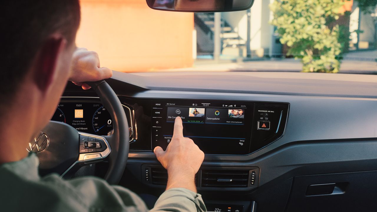 Muž ovláda internetový obchod VW upgrads vo VW Polo prostredníctvom displeja systému infotainment. Čiastočne vidieť volant a digitálny prístrojový panel. Cez čelné sklo vidieť strom a budovu.