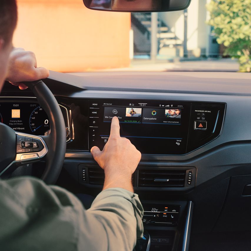 Muž ovláda internetový obchod VW upgrads vo VW Polo prostredníctvom displeja systému infotainment. Čiastočne vidieť volant a digitálny prístrojový panel. Cez čelné sklo vidieť strom a budovu.
