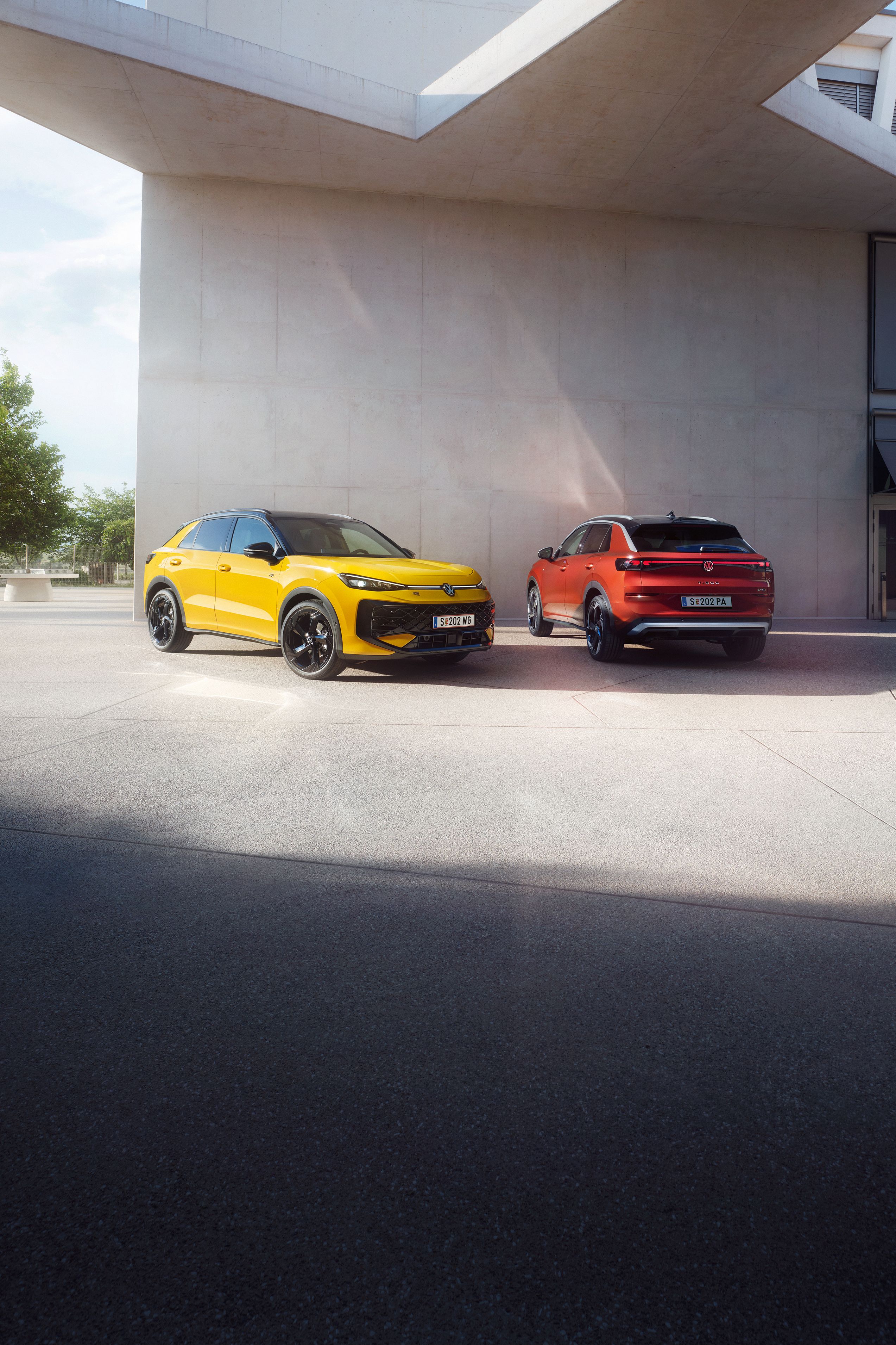 Žltý VW T-Roc R-Line a červený VW T-Roc Style stoja vedľa seba pred modernou budovou.