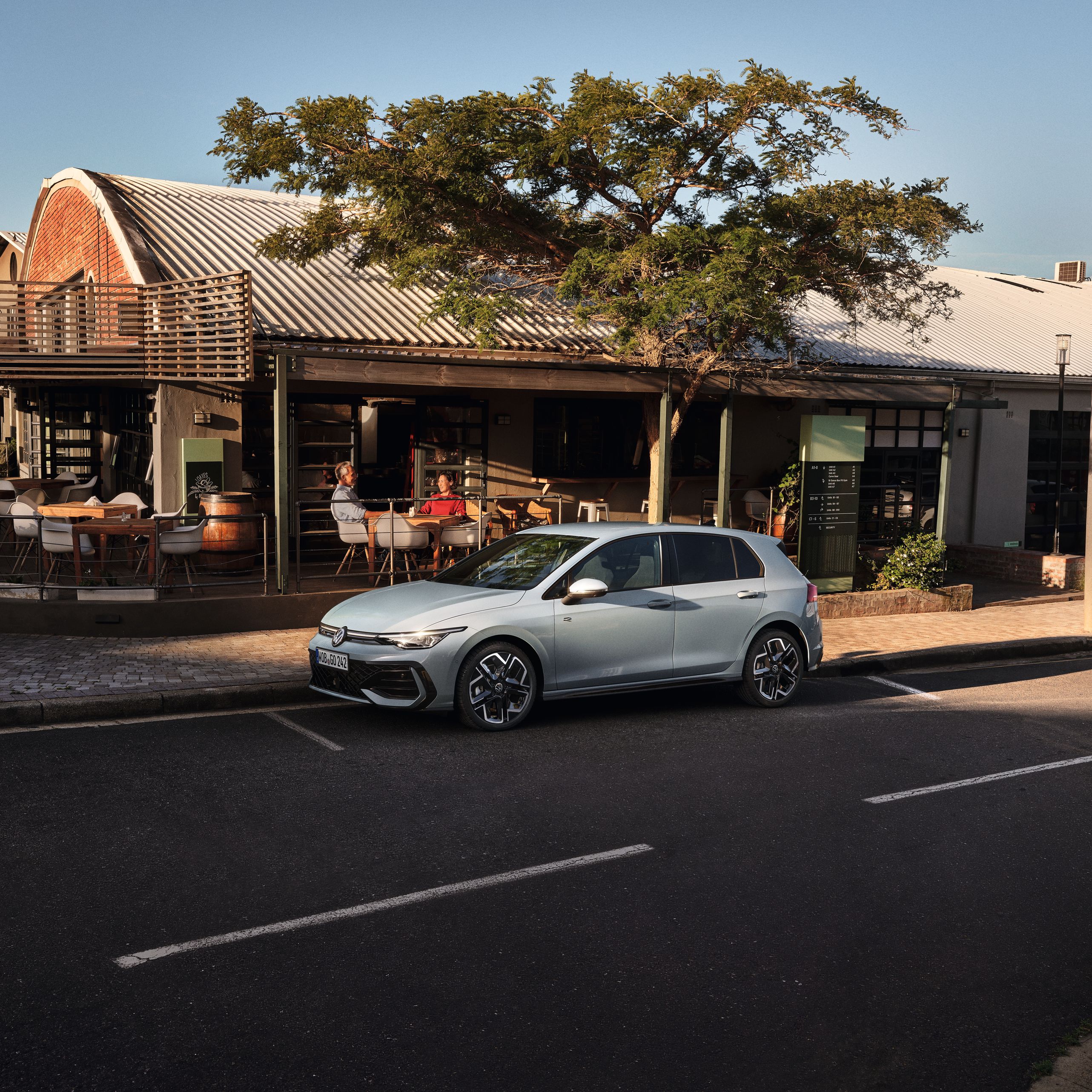 Biely Golf R-Line zaparkovaný na parkovacom mieste na ceste pred reštauráciou. Za vozidlom je reštaurácia a na terase sedia zákazníci.