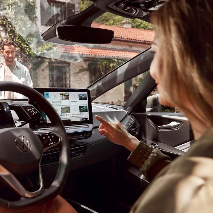 Interiér moderného vozidla s digitálnymi displejmi a dotykovým ovládaním. Žena sedí na sedadle vodiča a ovláda funkcie systému infotainment. Von pred vozidlom stojí muž a pozerá sa na ženu vo vozidle.