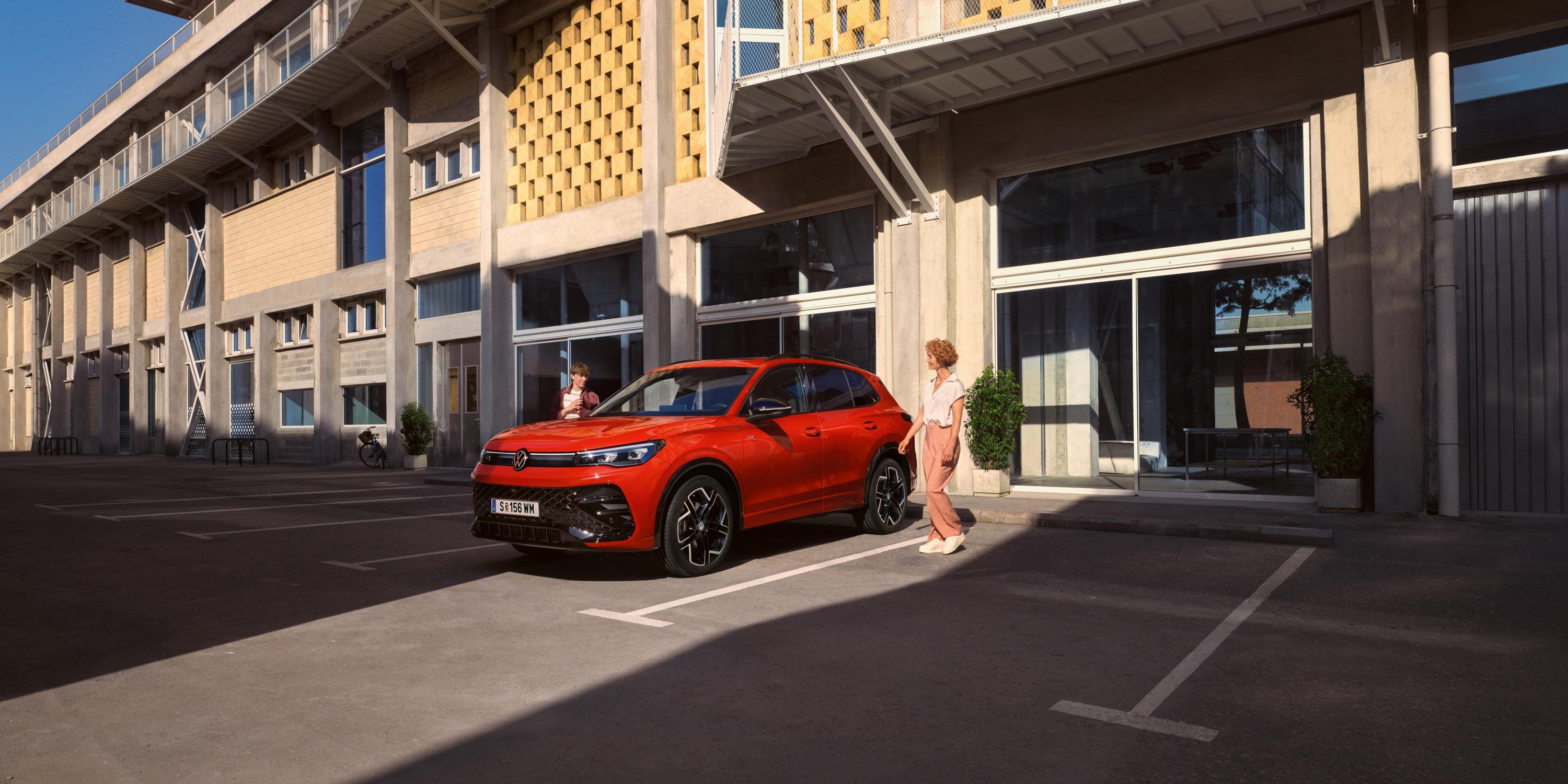 Červený VW Tiguan z bočného pohľadu stojí pred budovou, okolo prechádza žena.