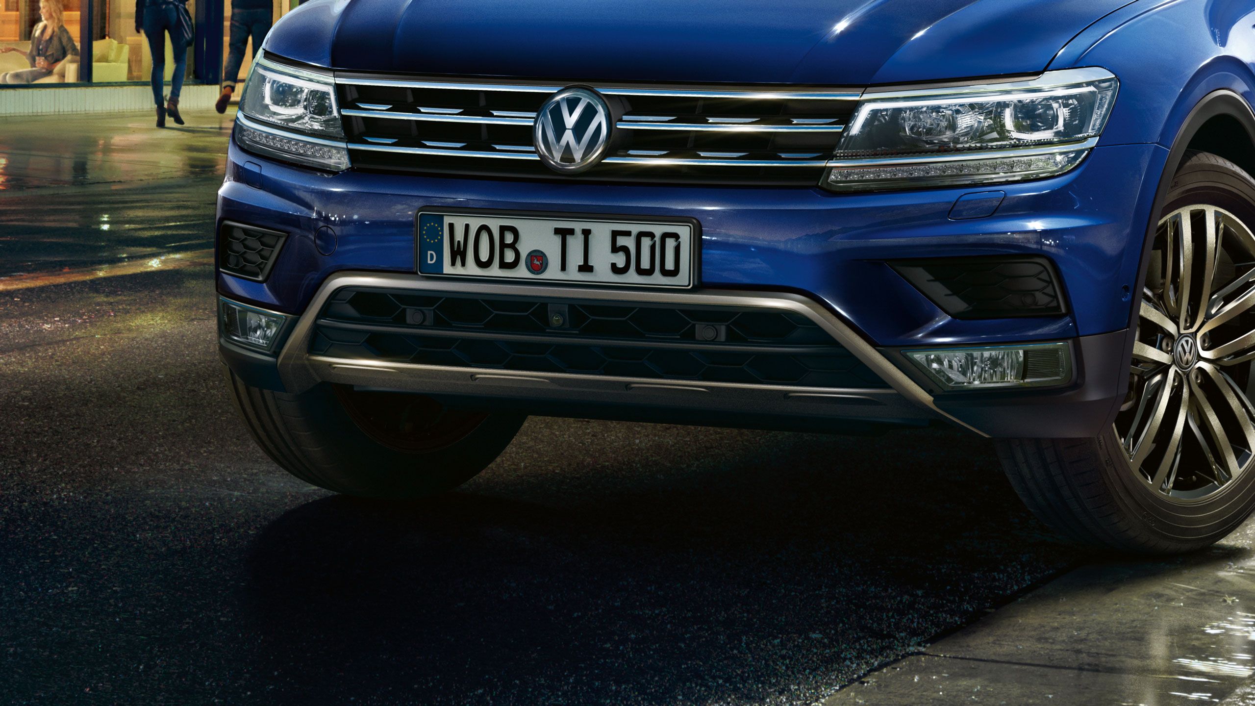 Modré SUV značky Volkswagen zaparkované na mokrej ceste. Na vozidle je vidieť predný nárazník a evidenčné číslo WOB TI 500. Na pozadí sú neostrí chodci a mestská  budova.