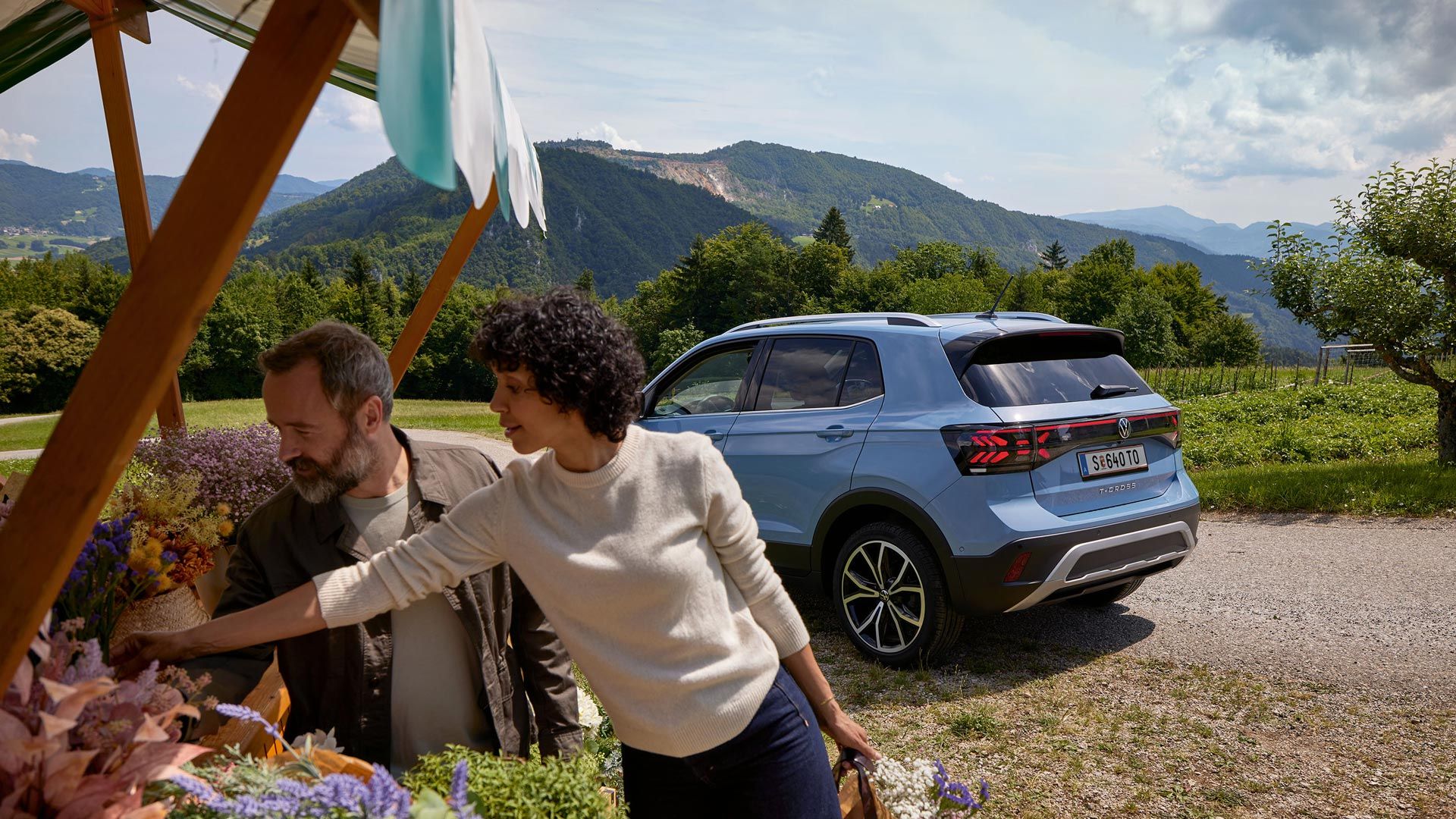 Muž a žena stoja pri kvetinovom stánku, za nimi modrý VW T-Cross pred hornatou krajinou.