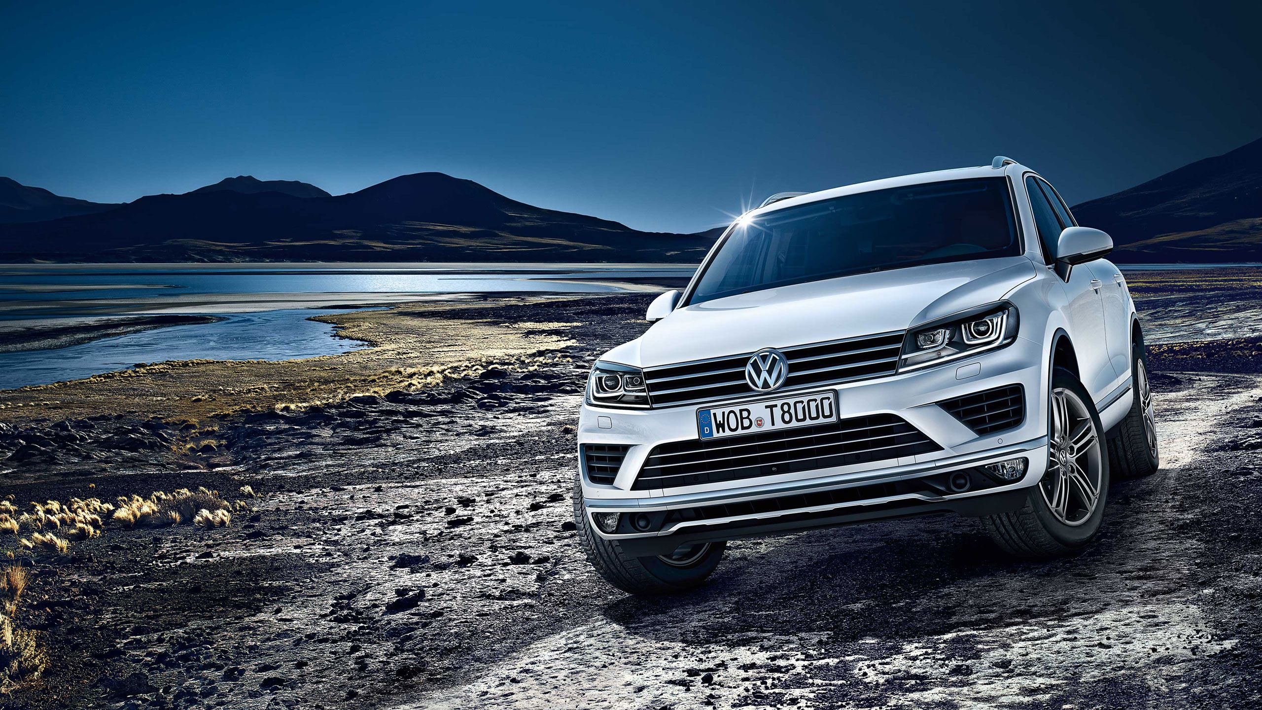 Biele SUV značky Volkswagen je zaparkované na kamenistej ceste pri vodnej ploche s pozadím hôr a jazera. Obsadenie je scénické a dramatické. Slnko jemne svieti na prednú časť auta a odráža sa v čelnom skle.