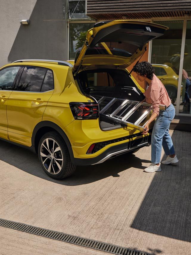 Mladá žena vkladá rebrík do batožinového priestoru žltého VW T-Cross. Vozidlo je zaparkované pred moderným rodinným domom s peknou záhradou s množstvom zelene.