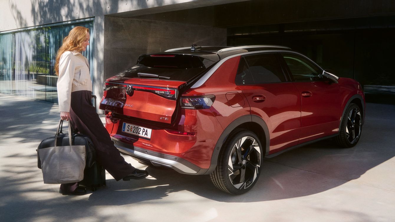 Žena s veľkou taškou stojí za červeným SUV T-Roc, ktoré je zaparkované pred modernou budovou a pohybom nohy otvára dvere batožinového priestoru.