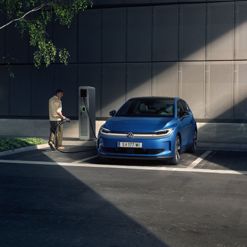 Modré ID. Polo stojí na parkovisku vedľa nabíjacej stanice. V pozadí je vidieť budovu a strom.