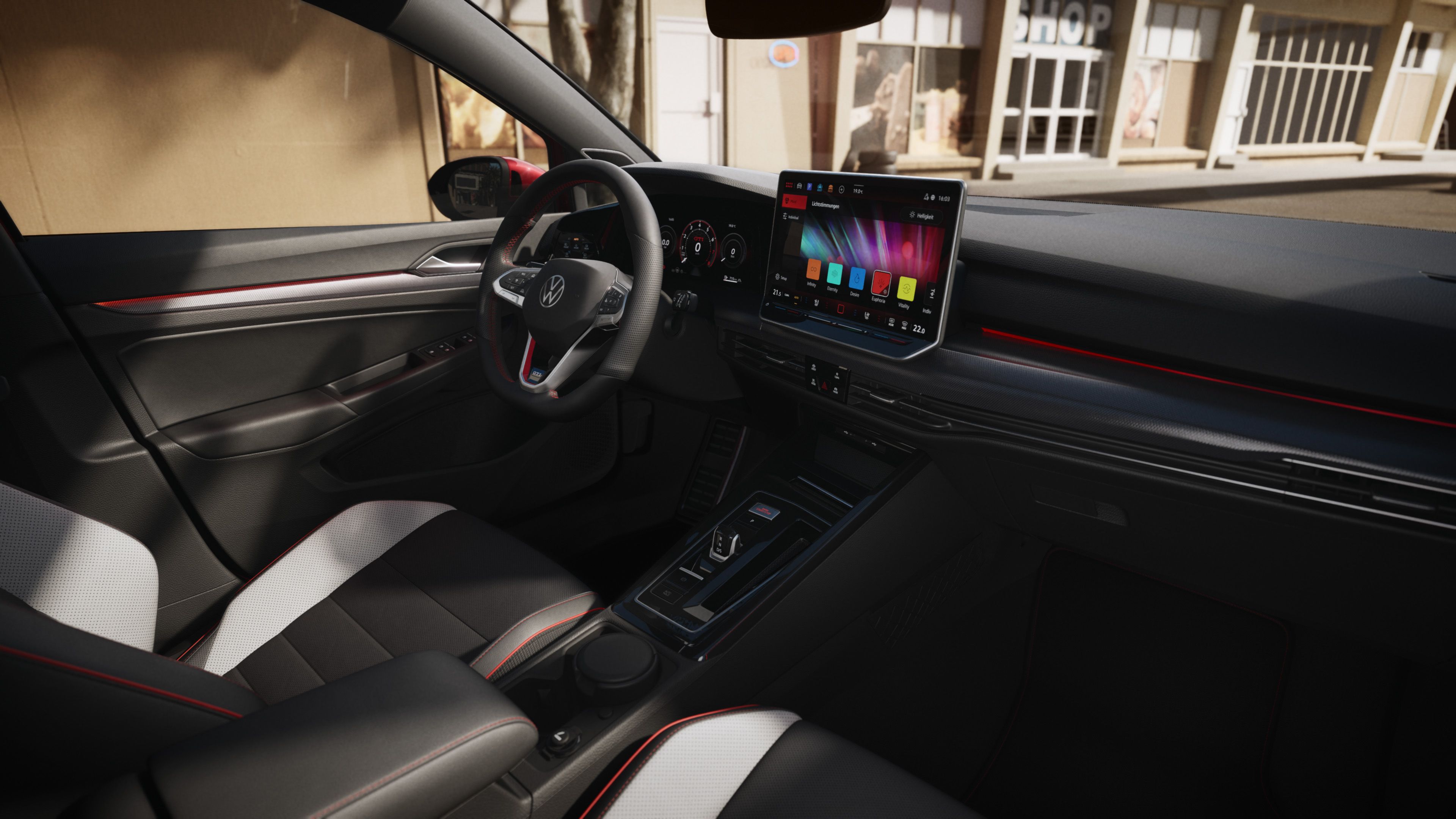 Pohľad do interiéru VW Golf GTI zo sedadla spolujazdca. Dobre viditeľné sedadlo vodiča v dvojfarebnom čierno-bielom čalúnení, volant, digitálny prístrojový panel, displej systému infotainment, palubná doska čiernej farby, obklad dverí. Cez čelné sklo vidieť svetlú budovu.