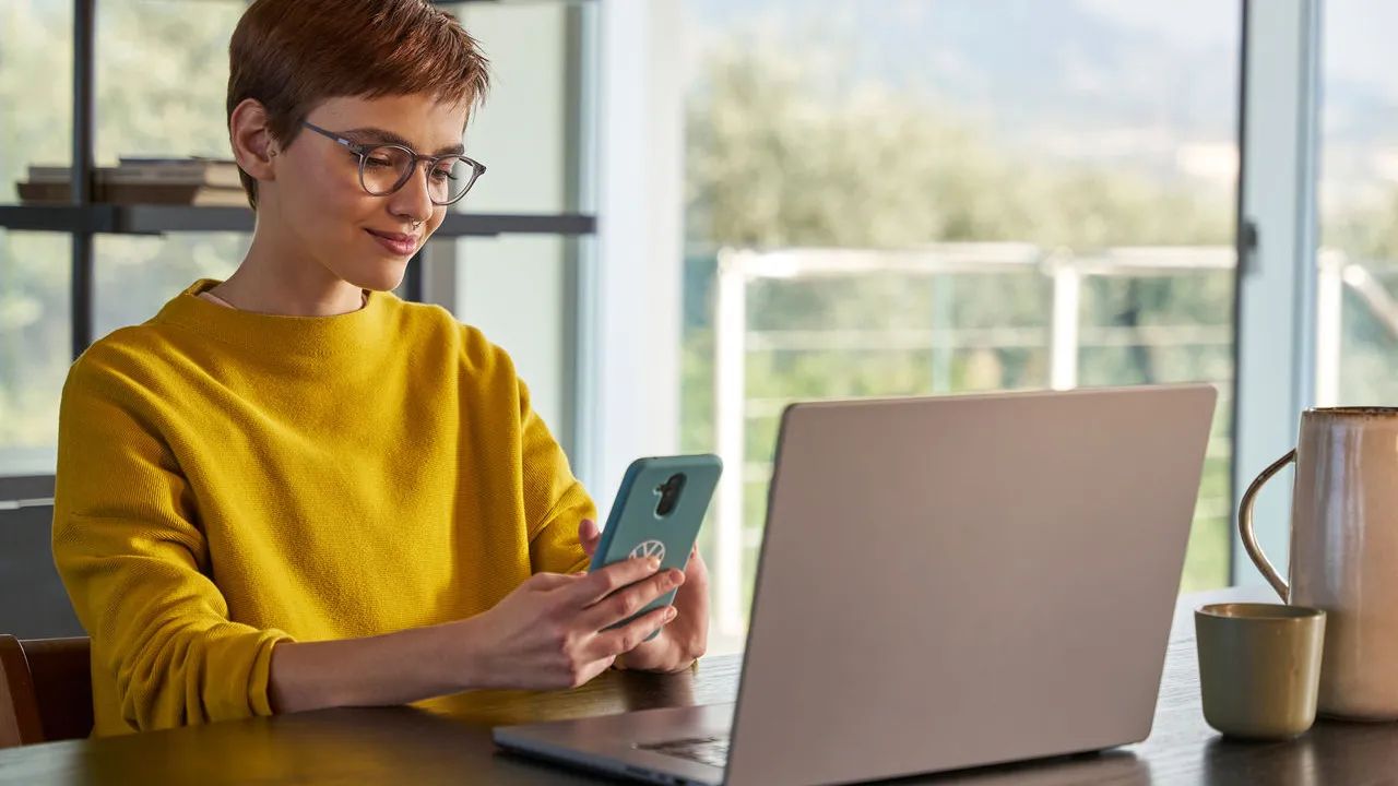 Žena sa pozerá na svoj mobilný telefón pred svojím notebookom.