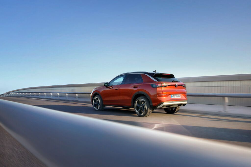 Červený SUV automobil jazdí po modernom moste. Vozidlo má športový dizajn a tmavé zadné okná. Obloha je jasná a modrá.