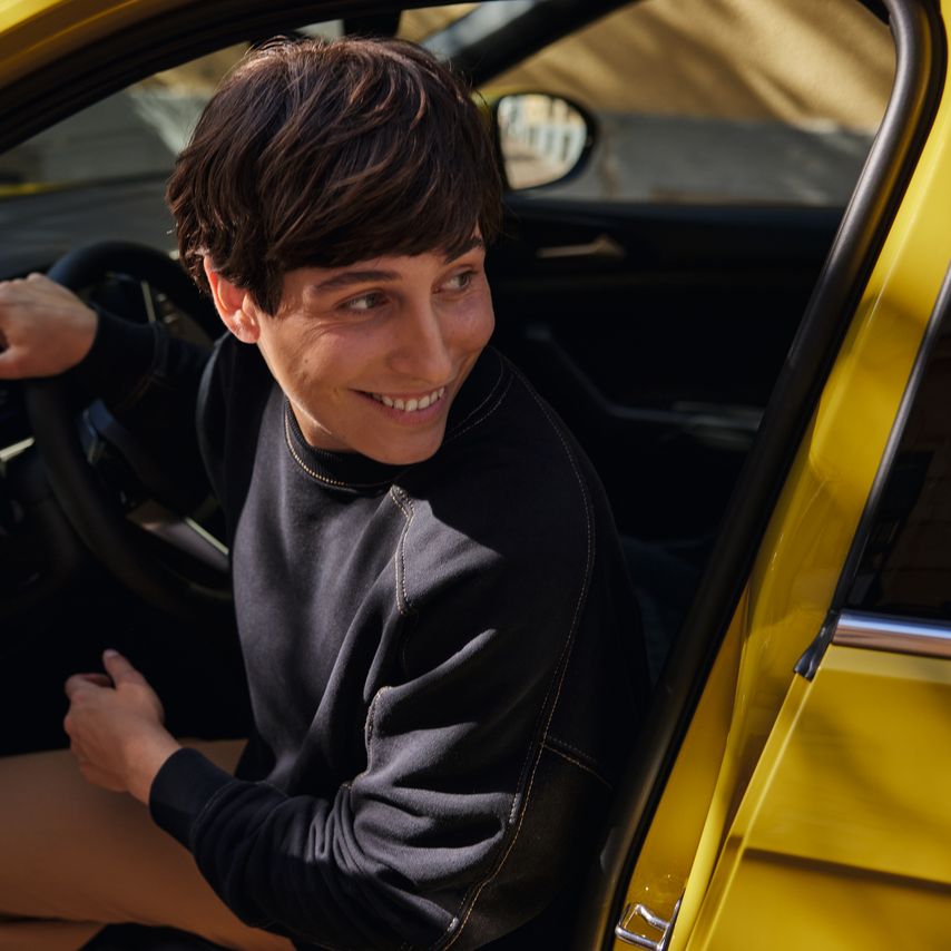 Mladá žena na sedadle vodiča žltého VW T-Cross sa s úsmevom pozerá cez otvorené dvere.