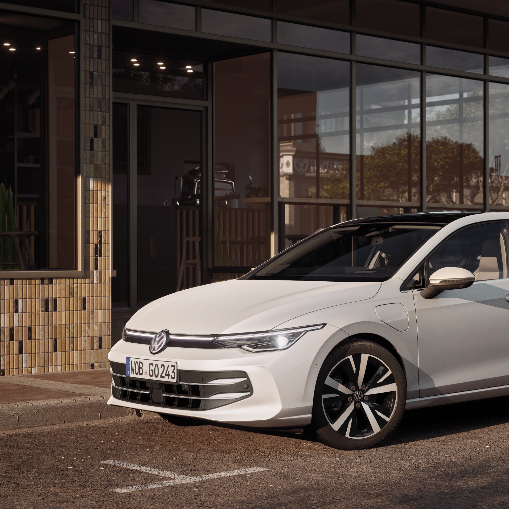 Biely hatchback Volkswagen zaparkovaný pred modernou budovou s čiastočne sklenenou fasádou. Auto stojí na slnečnej ulici s dláždeným chodníkom. Vozidlo má elegantný dizajn a športové disky z ľahkej zliatiny.