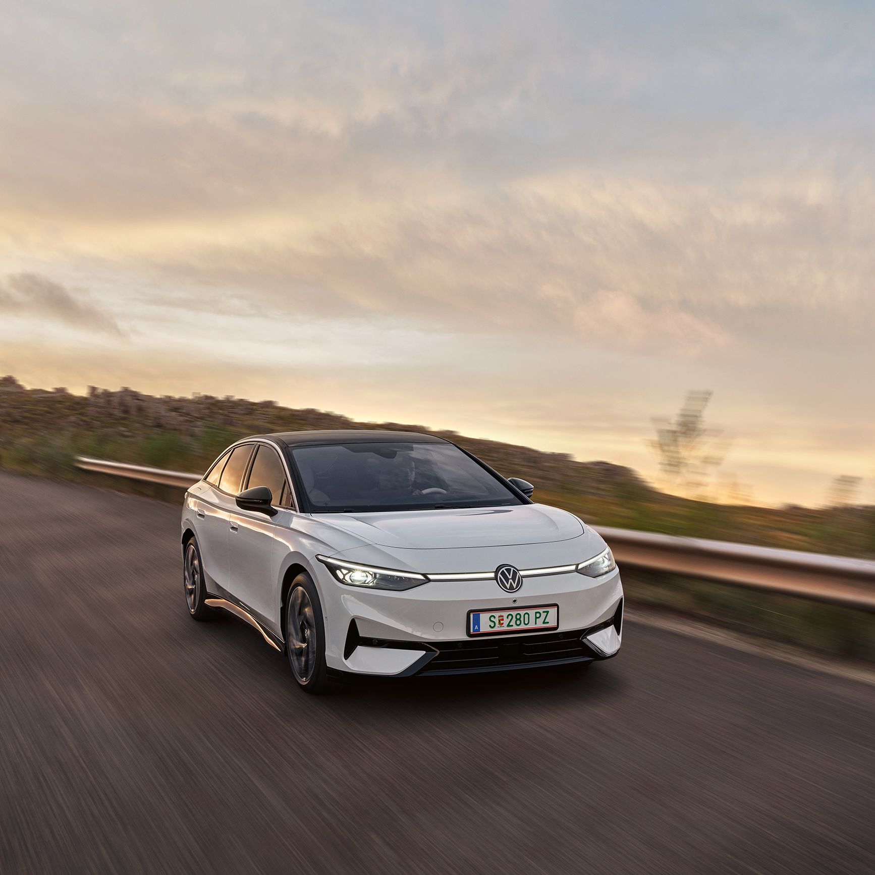 Biely elektrický Volkswagen ID.7 jazdí po ceste pri západe slnka. Rozmazané pozadie naznačuje pohyb. Auto je elegantné a moderné.