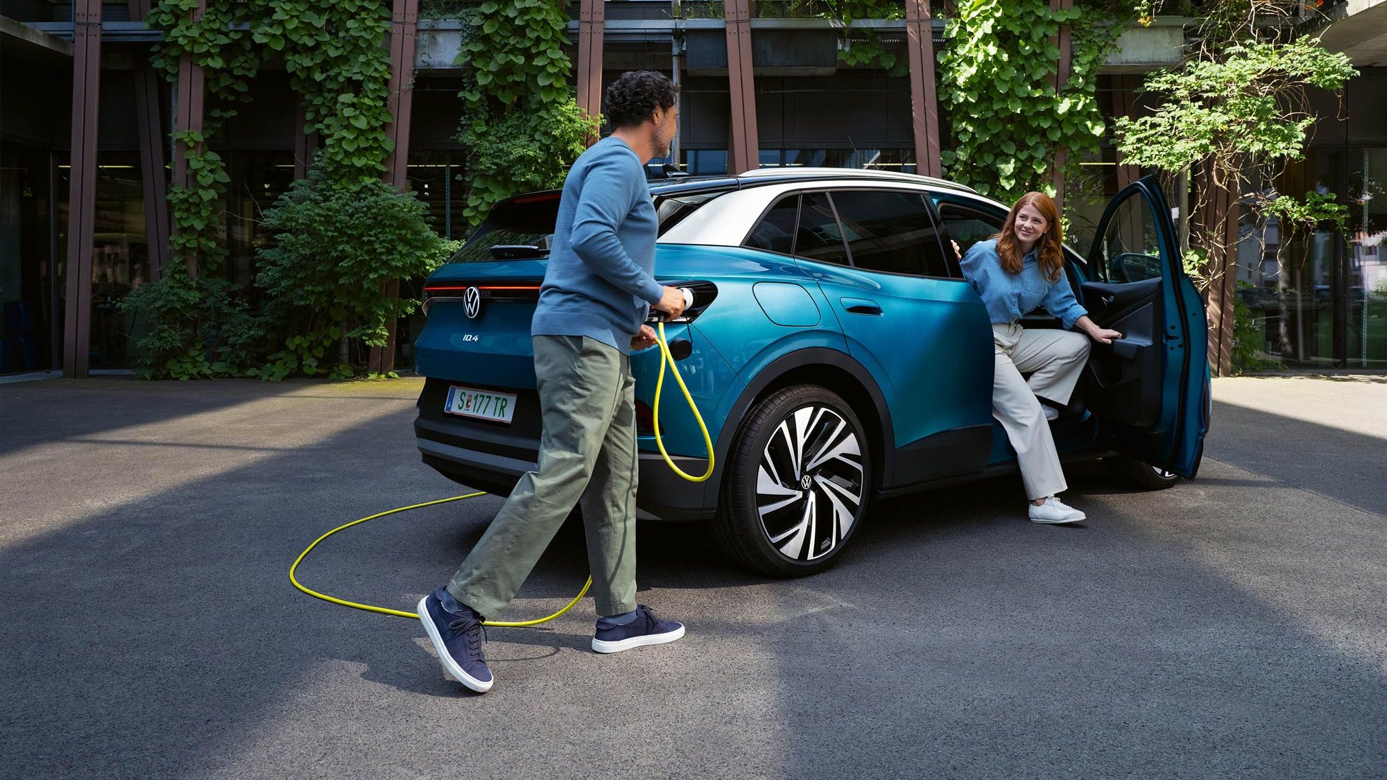 Muž so žltým nabíjacím káblom kráča k zadnej časti zaparkovaného modrého vozidla VW ID.4. Z dverí spolujazdca vystupuje mladá žena. vozidlo stojí pred modernou budovou, ktorá je obrastená zeleňou.