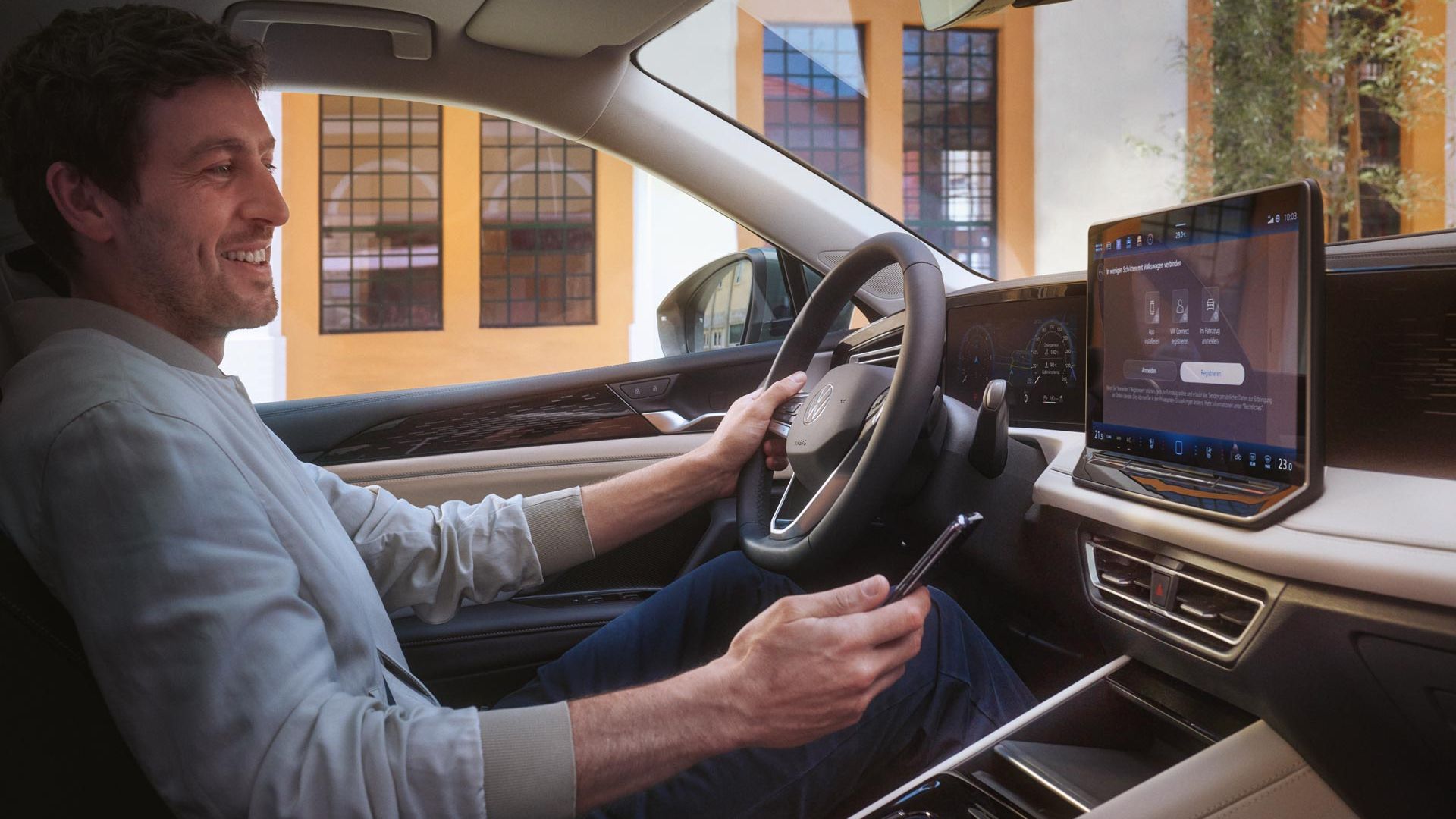 Muž sedí na mieste vodiča jednou rukou drží volant, druhou smartfón, ktorým sa spája so systémom vozidla. Za vozidlom je moderná budova.