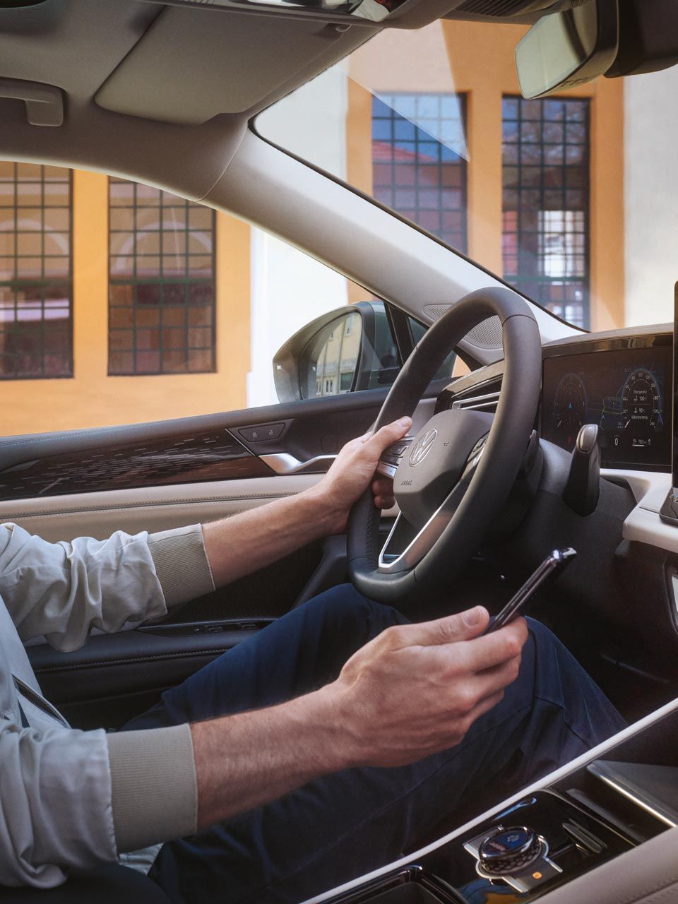 Muž sedí na mieste vodiča jednou rukou drží volant, druhou smartfón, ktorým sa spája so systémom vozidla. Za vozidlom je moderná budova.