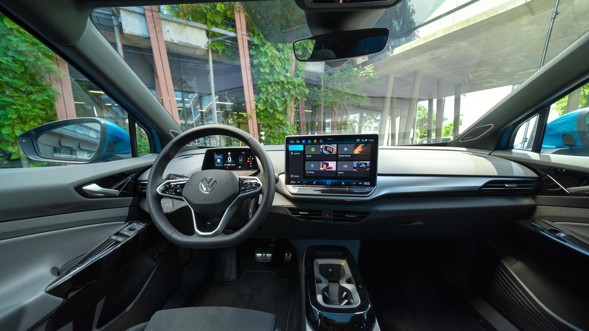 Interiér moderného Volkswagen ID.4. Na obrázku je elegantná palubná doska s integrovaným dotykovým displejom. Táto obrazovka zobrazuje rôzne aplikácie a informácie. Vidieť tiež časť predných sedadiel, digitálny prístrojový panel, stredovú konzolu a obklad dverí v modernom dizajne. Cez čelné sklo vidieť pohľad na modernú stavbu.