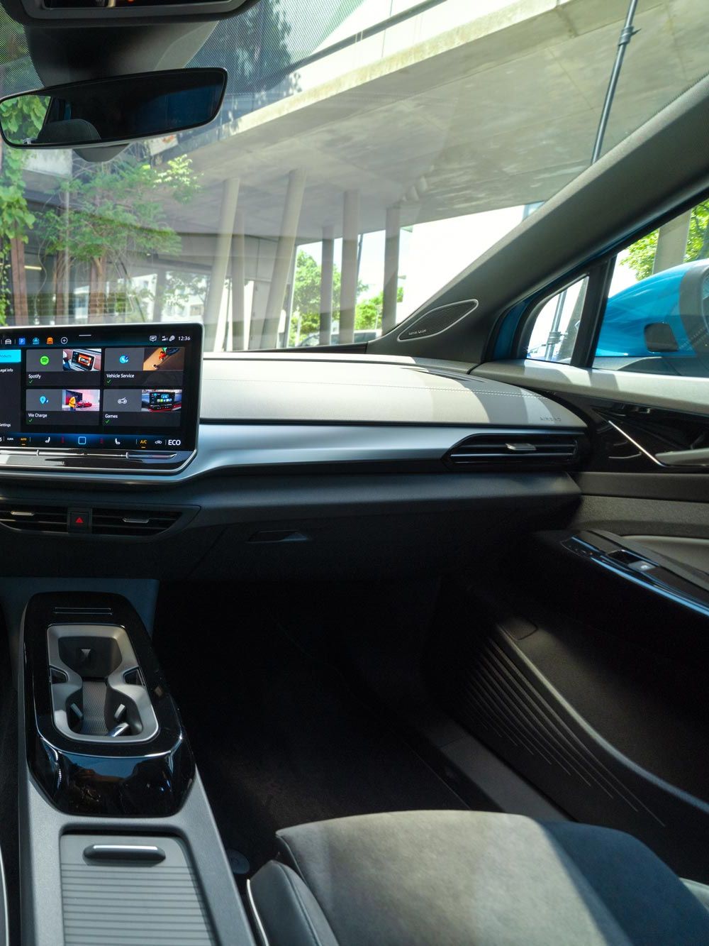 Interiér moderného Volkswagen ID.4. Na obrázku je elegantná palubná doska s integrovaným dotykovým displejom. Táto obrazovka zobrazuje rôzne aplikácie a informácie. Vidieť tiež časť predných sedadiel, digitálny prístrojový panel, stredovú konzolu a obklad dverí v modernom dizajne. Cez čelné sklo vidieť pohľad na modernú stavbu.